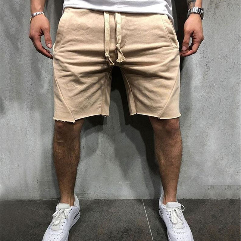 Men's beige cotton gym shorts with drawstring and raw hem, worn with white sneakers against gray wall