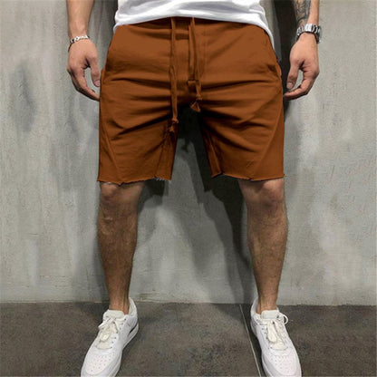 man wearing rust orange cotton gym shorts with drawstring, white sneakers, standing against gray concrete wall