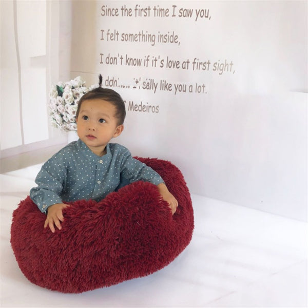 Child sitting in a soft, fluffy red round cushion pet bed in a bright room with wall text and flowers