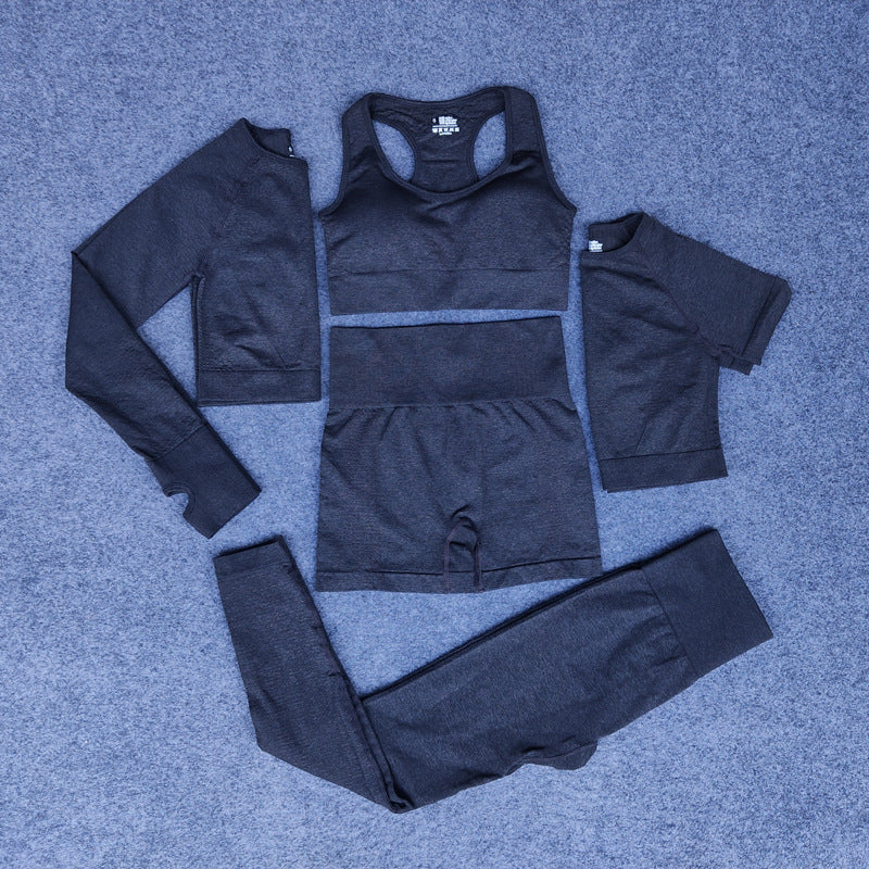 Charcoal gray seamless four-piece activewear set including long sleeve crop top, sports bra, shorts, and leggings on gray carpet