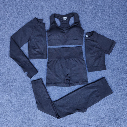 Charcoal gray seamless four-piece activewear set including long sleeve crop top, sports bra, shorts, and leggings on gray carpet