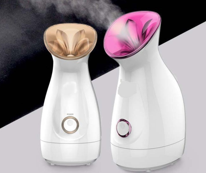 Two white nano ion facial steamers with gold and pink nozzles emitting steam on black and gray background
