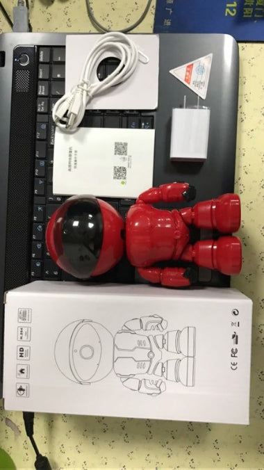 Red robot-shaped smart home security camera with USB cable, charger, manual, and packaging on laptop keyboard