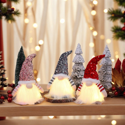 Three lit Christmas gnome decorations with sequined hats in pink, gray, and red on wooden surface with snowy miniature trees