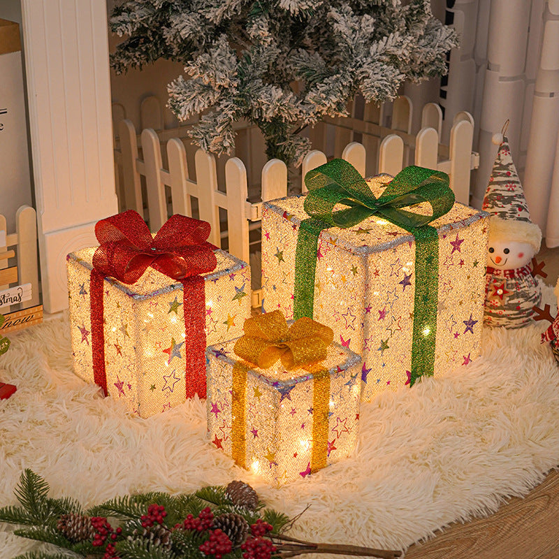 Three illuminated Christmas gift boxes with red, green, and gold bows on a white rug under a snowy pine tree