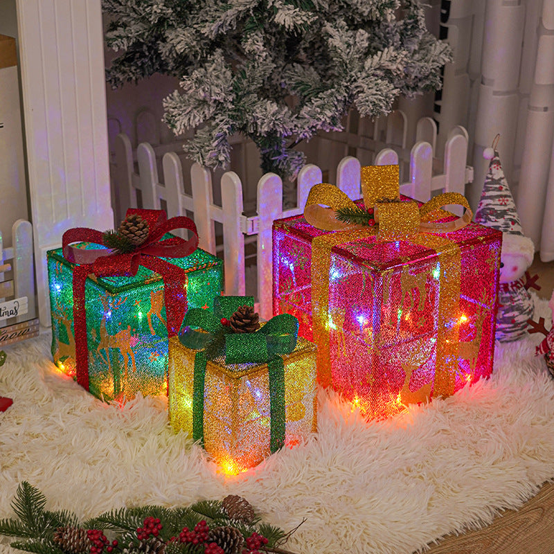 Three LED-lit Christmas gift boxes with glittery bows and pinecone accents by a white fence and frosted tree