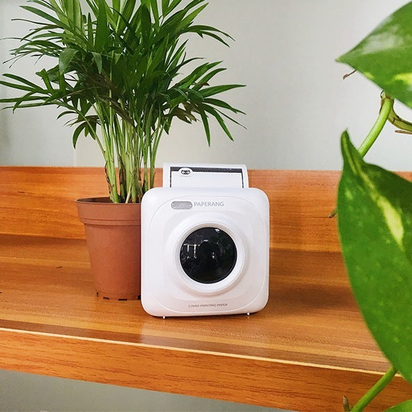 White portable Paperang thermal printer printing photo on wooden shelf next to potted plant
