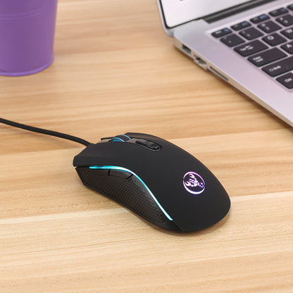Black wired gaming mouse with RGB lighting on wooden desk next to laptop and purple cup