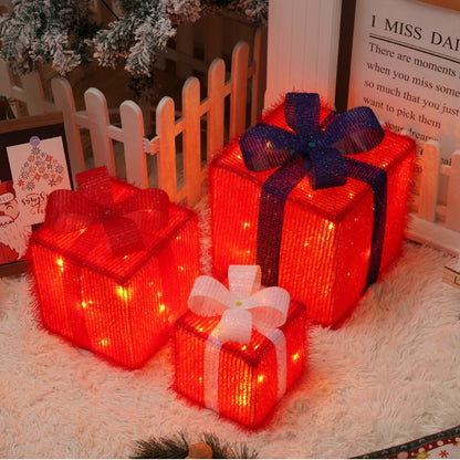 Three illuminated red Christmas gift boxes with bows in white, red, and blue on a white carpet by a white picket fence