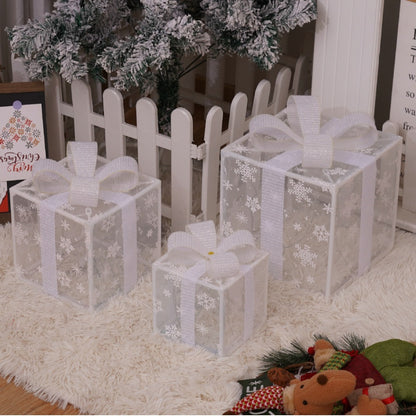 Three transparent Christmas gift boxes with white snowflake patterns and bows under a snowy tree near white picket fence on white rug
