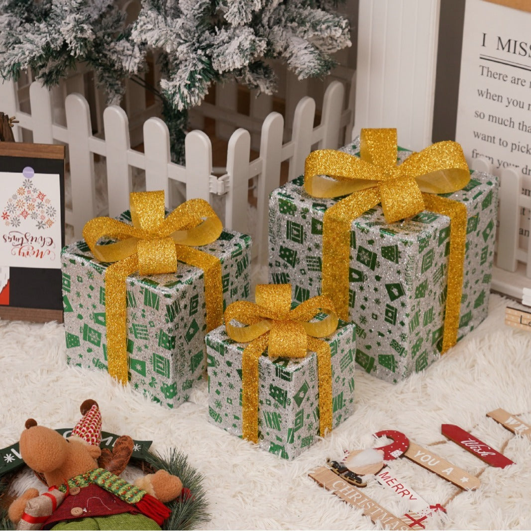 Three glittery Christmas gift boxes with gold ribbons under a snowy tree and holiday decor
