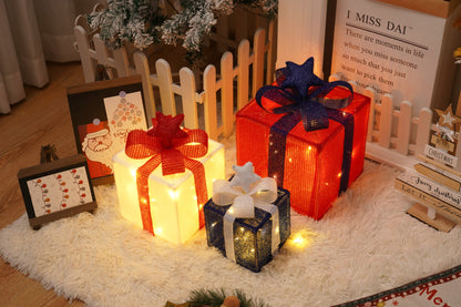 Three colorful Christmas gift box decorations with glowing lights on a white rug near festive holiday signs and a small wooden fence