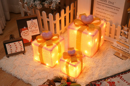Three glowing Christmas gift box decorations with golden ribbons and pink heart toppers on a furry white rug