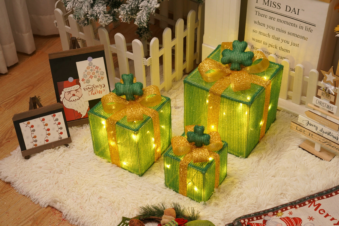 Three green Christmas gift boxes with golden ribbons and lights on a white rug near holiday decorations and a small white fence