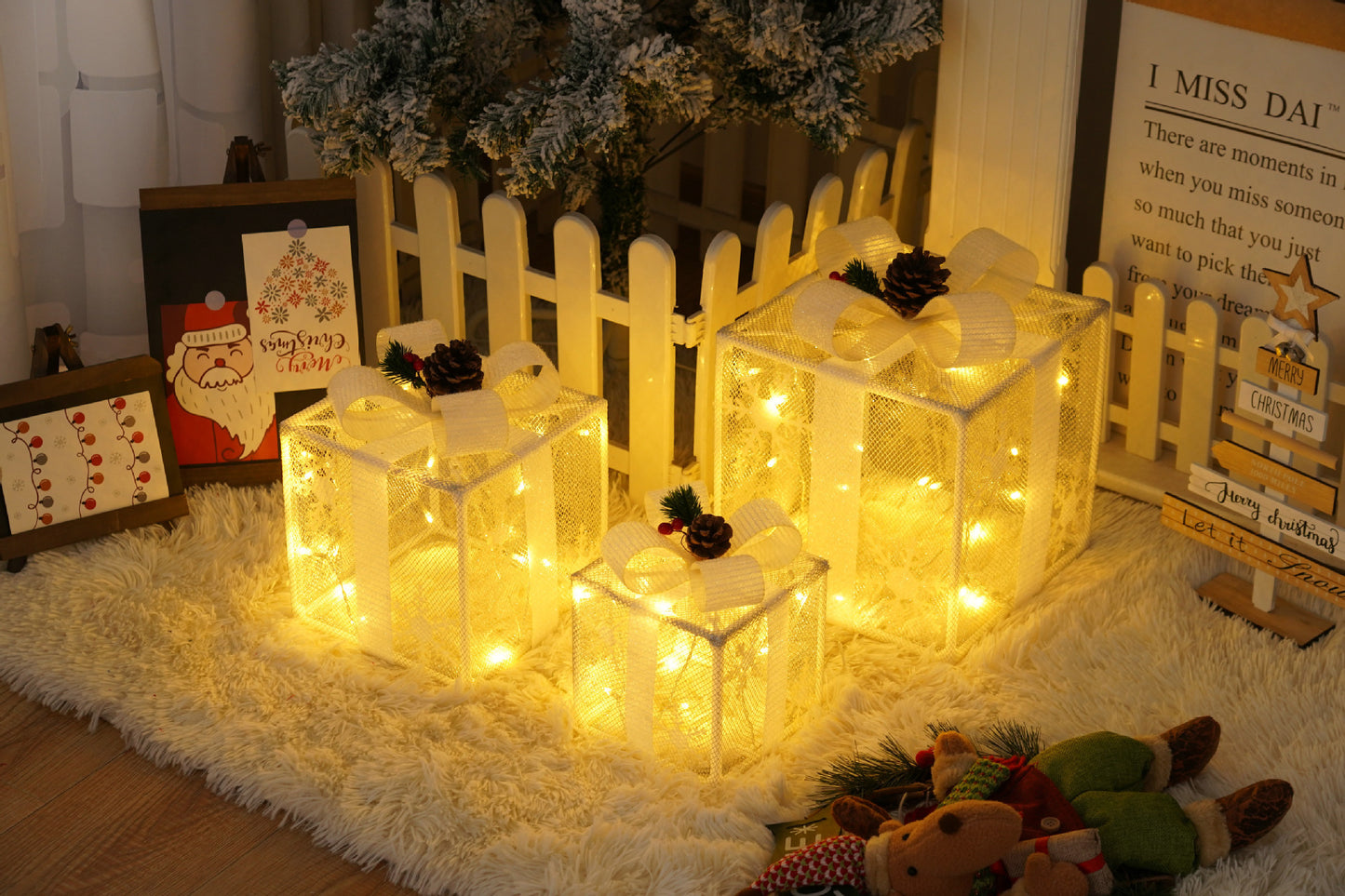 Three glowing Christmas gift box decorations with white ribbons and pinecones on a soft rug near a white picket fence and holiday signs