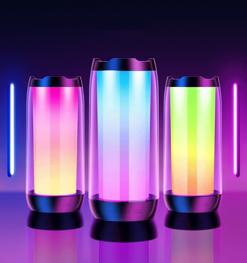 Three ambient light Bluetooth speakers with vibrant gradient colors on a reflective surface in a dark gaming room