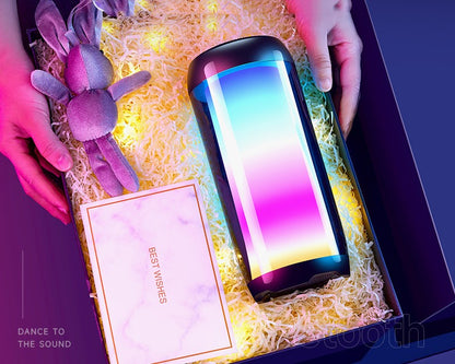 Hands holding a gift box with colorful ambient Bluetooth speaker, plush toy, and best wishes card on yellow shredded paper