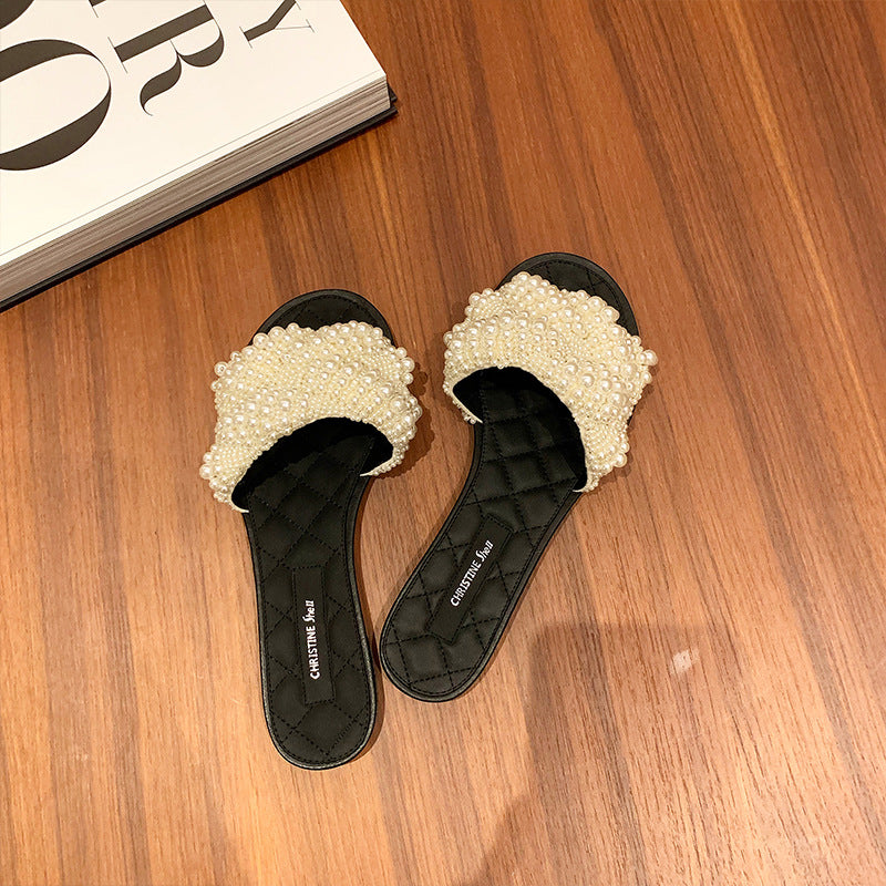 Black flat slide sandals with pearl-embellished wide straps on wooden floor next to a magazine