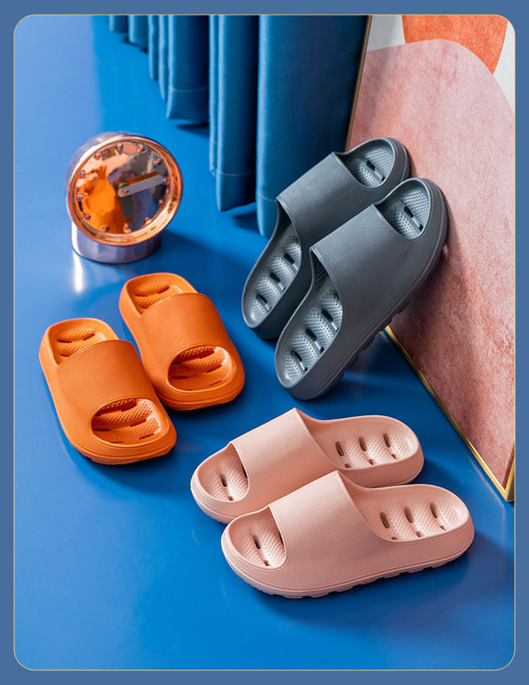 Three pairs of cushioned EVA slide slippers in orange, gray, and pink on a blue surface near a clock and curtain