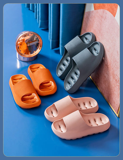 Three pairs of cushioned EVA slide slippers in orange, gray, and pink on a blue surface near a clock and curtain