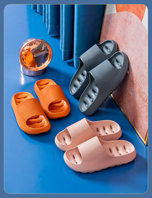 Three pairs of cushioned EVA slide slippers in orange, gray, and pink on a blue surface near a clock and curtain