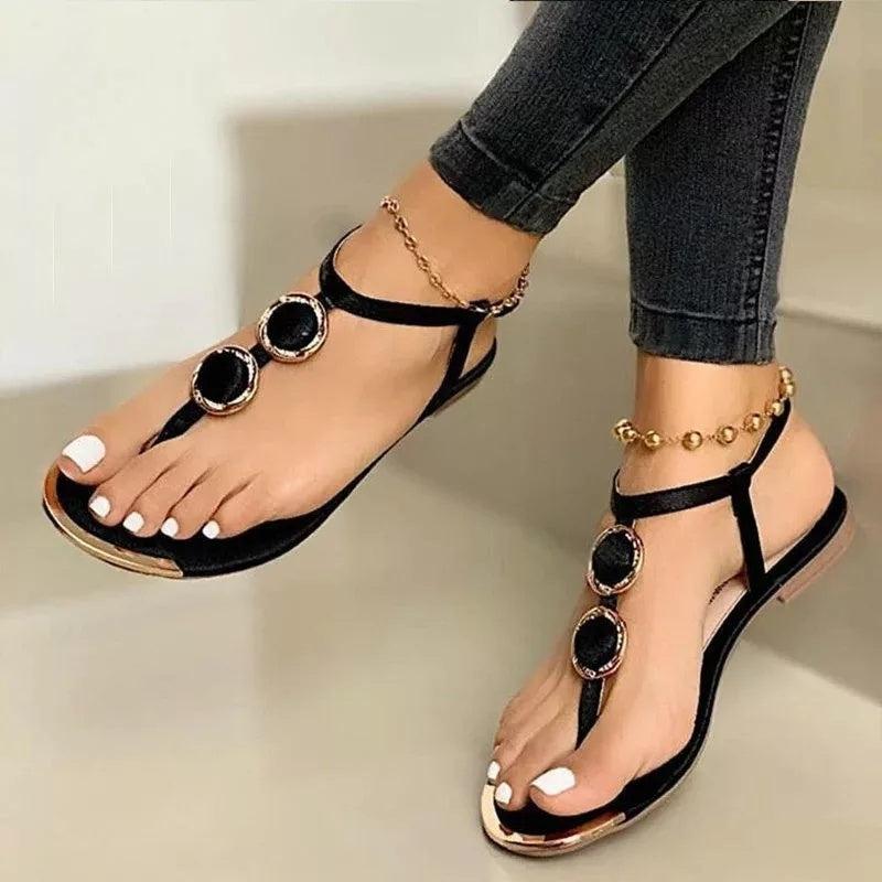 Women's black flat sandals with circular metal buckles, worn with dark jeans and gold anklets