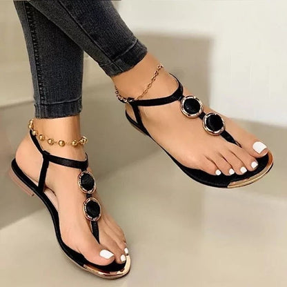 Women's black flat sandals with metal buckle accents and gold anklet on feet with white nail polish