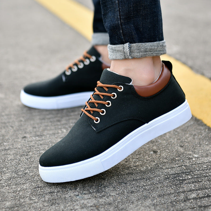 Black men's casual sneakers with brown laces and heel collar, white rubber sole, worn with cuffed jeans on asphalt