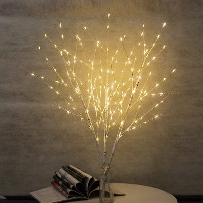 White birch tree branches wrapped with warm white LED lights in a glass vase on a table with books