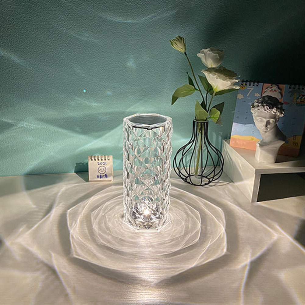 Clear crystal table lamp casting decorative light patterns on white surface with flowers and a bust nearby