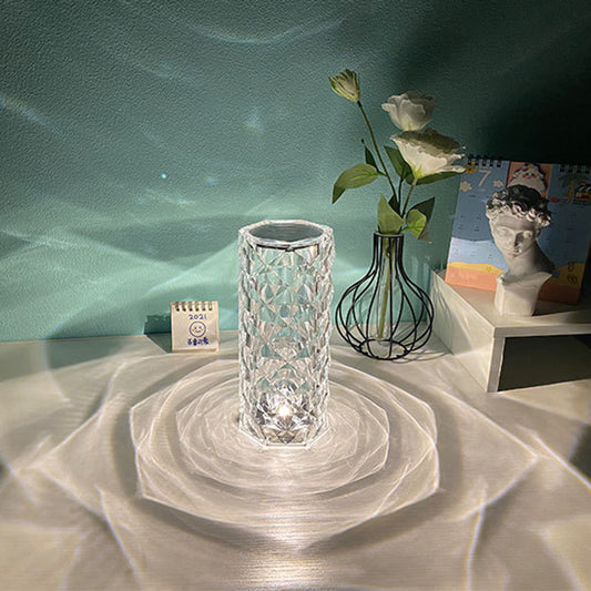 Clear crystal table lamp casting decorative light patterns on white surface with flowers and a bust nearby