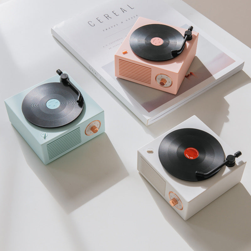 Three retro mini phonograph Bluetooth speakers in pastel pink, blue, and white on white surface with magazine