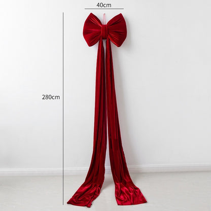 Large wine red velvet Christmas bow decoration with long trailing ribbons against white wall
