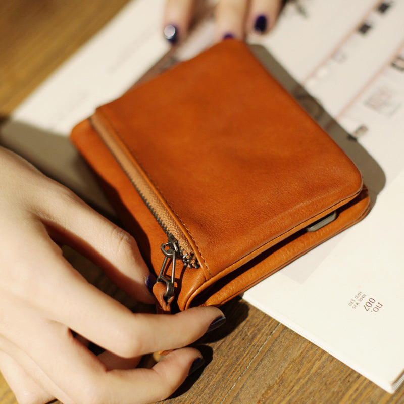 Slim bifold brown leather wallet with zipper, held by hand on wooden table with open book