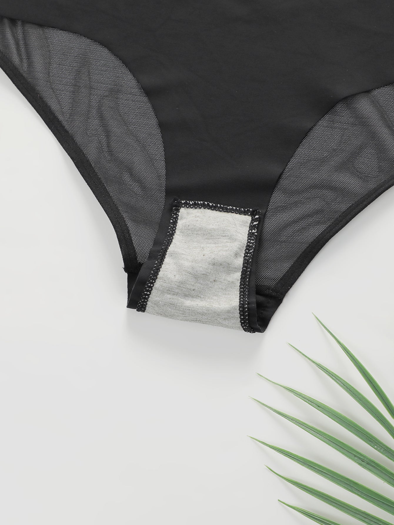 Close-up of black women's lingerie panties with sheer mesh panels and grey cotton gusset on white surface with green plant