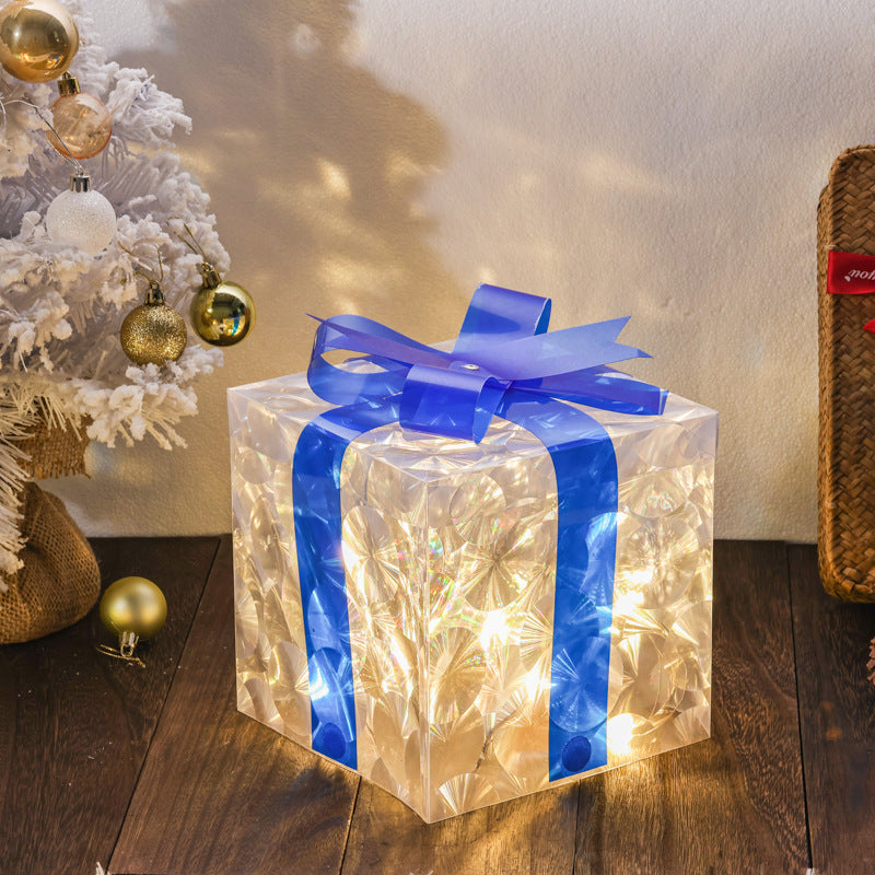 Glowing luminous Christmas gift box with blue ribbon on wooden floor near decorated white Christmas tree