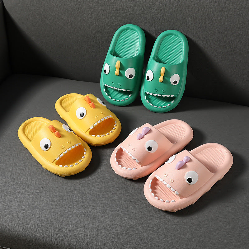 Three pairs of kids shark-themed slippers in yellow, green, and pink with non-slip soles on a gray surface