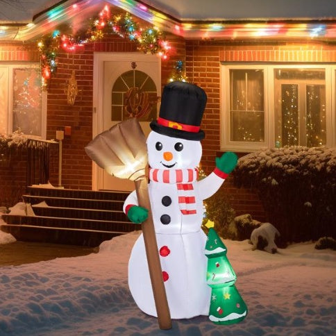 Inflatable LED snowman with top hat, scarf, broom and Christmas tree decoration outside a lit brick house in snow