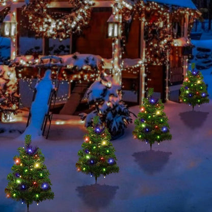 Four small solar-powered Christmas trees with multicolor LED lights on snowy ground near a decorated house
