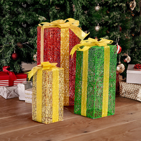 Three tall lighted gift boxes in red, green, and gold with yellow ribbons under a decorated Christmas tree