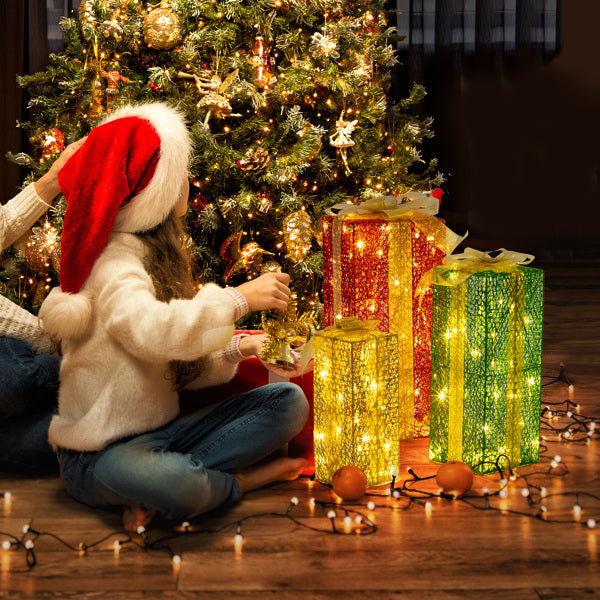 Child in Santa hat decorating Christmas tree near three lighted gift box decorations in red, yellow, and green