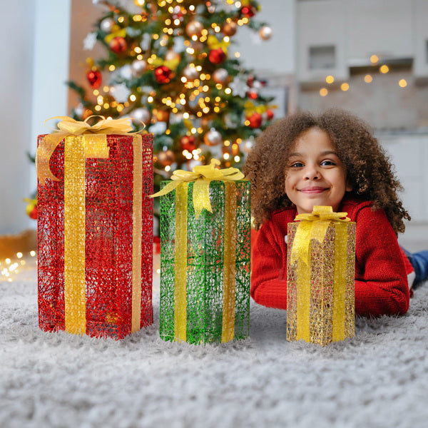Child in red sweater lying on carpet with three lighted decorative gift boxes in red, green, and gold near a decorated Christmas tree