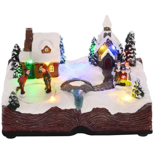 Animated Christmas village scene with snowy cottages, lit trees, figurines, and an open book base