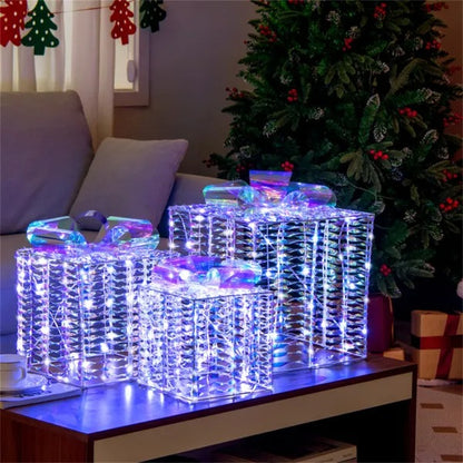 Iridescent lighted Christmas gift box decorations in three sizes on a table near a decorated Christmas tree
