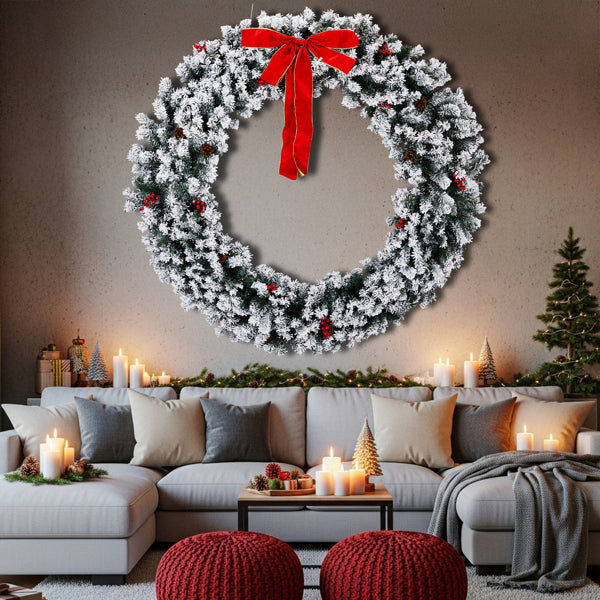 Large snow-flocked Christmas wreath with red bow and berries above cozy living room with candles and festive decor