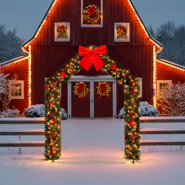 Lighted Christmas garland archway with red bow and decorations in snowy yard in front of a lit red barn with wreaths