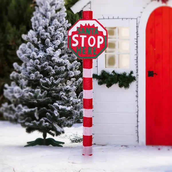 Lighted Christmas yard sign reading 'Santa Stop Here' in red and white by snowy tree and white house with red door