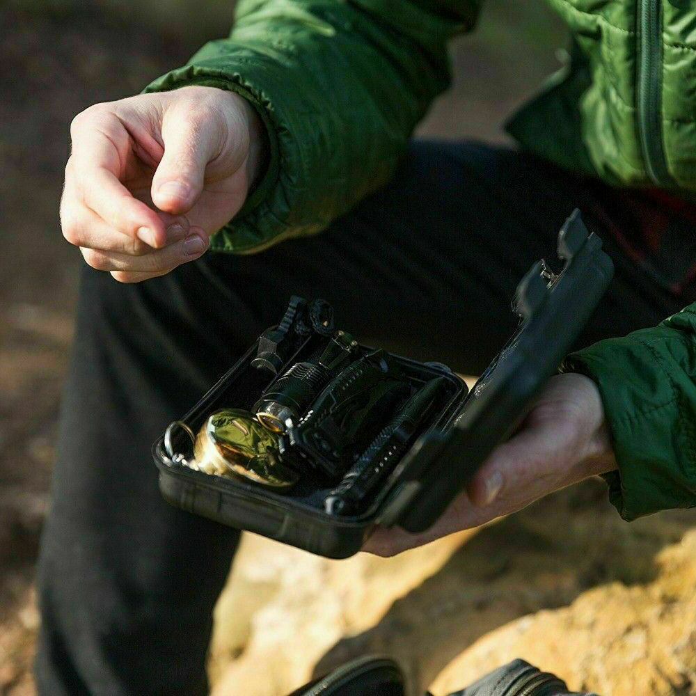 Person in green jacket holding open black survival kit with flashlight and compass outdoors