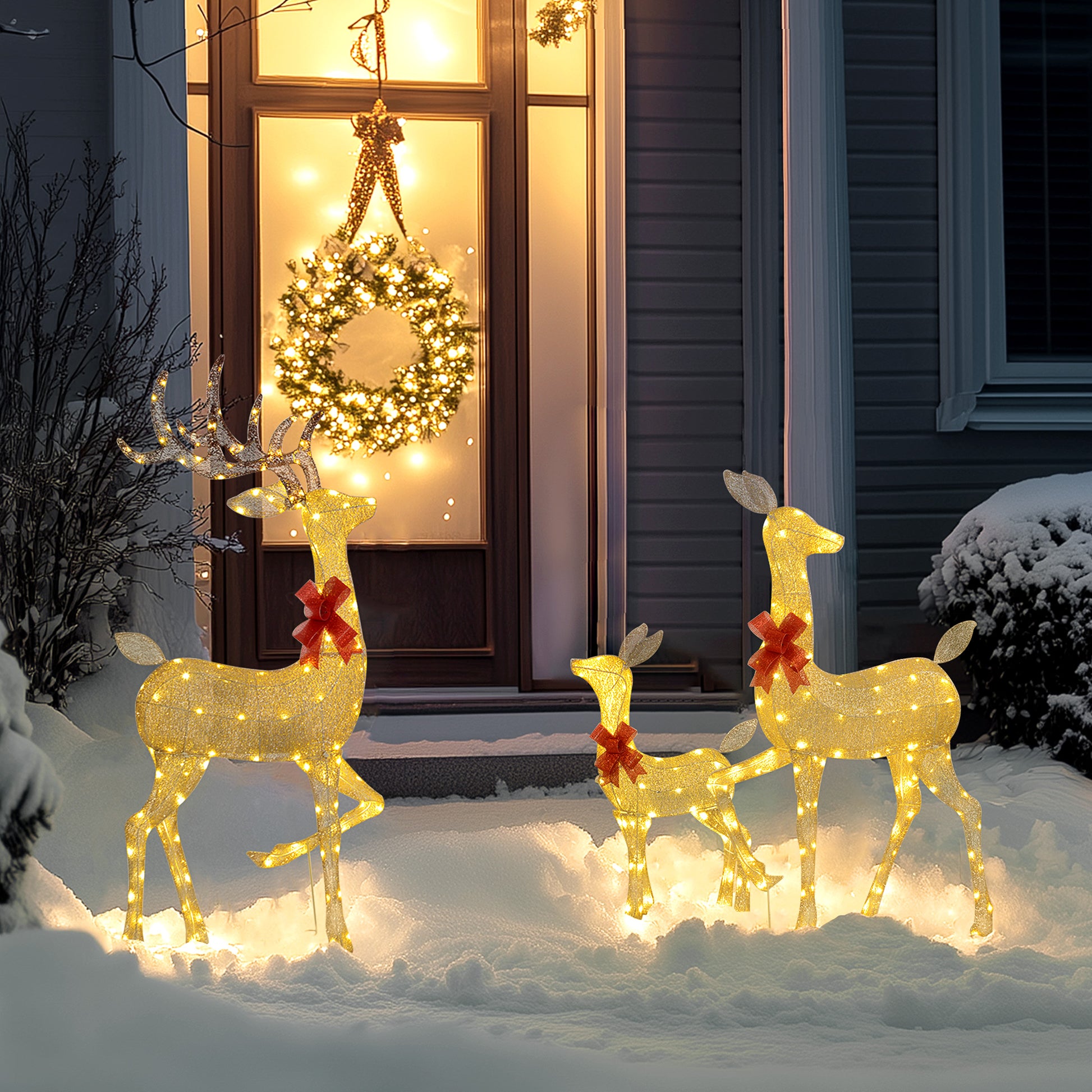 Three warm white LED-lit reindeer decorations with red bows in snowy front yard near illuminated Christmas wreath on door