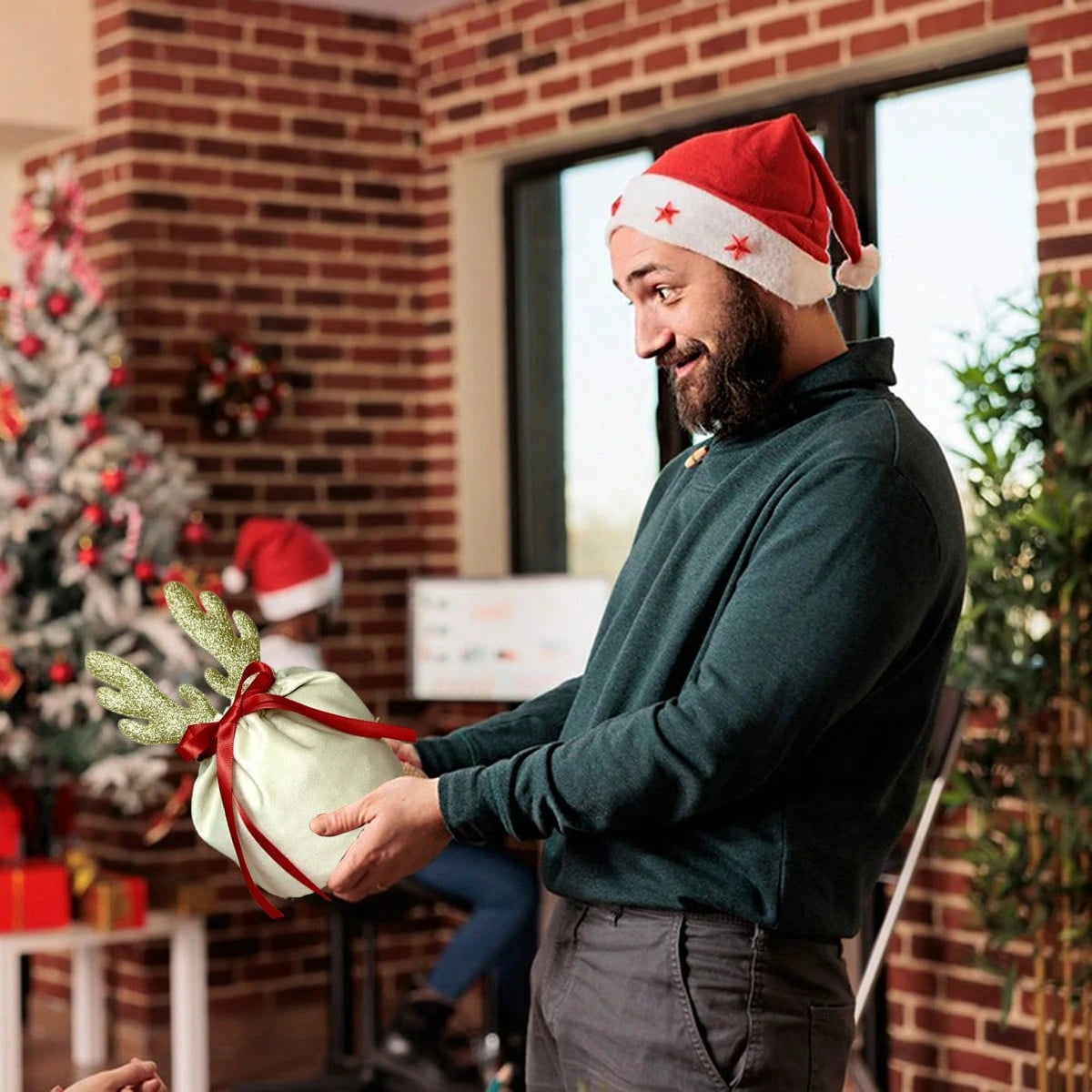 Man in Santa hat holding Christmas gift bag with gold antlers and red ribbon in decorated room